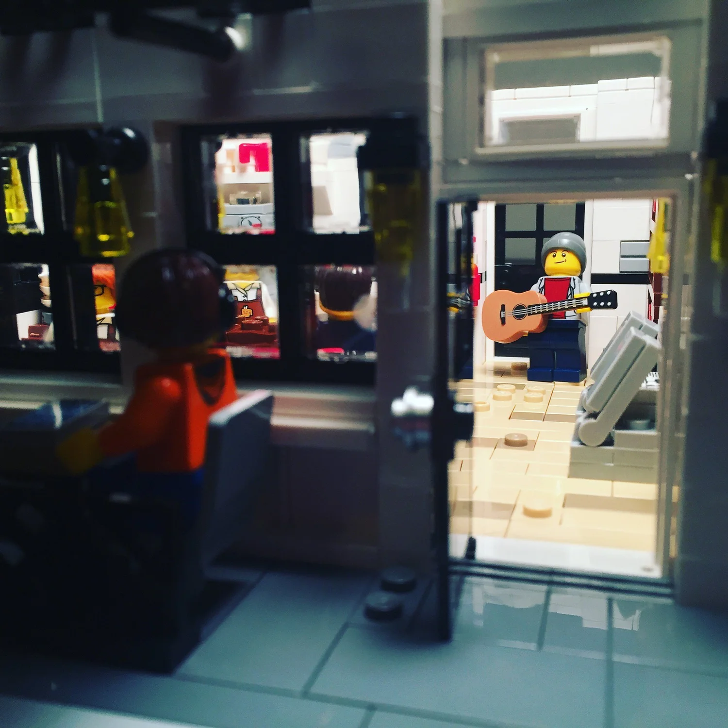 Songwriter minifigure with an acoustic guitar seen through the café door, with a patron in the foreground
