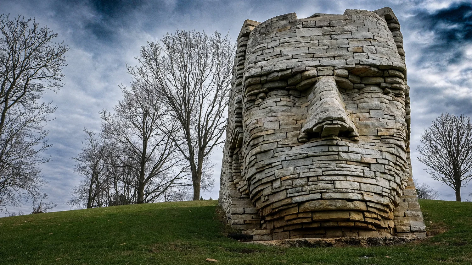 Weathered stacked-stone head sculpture on a grassy hill beneath a cloudy sky
