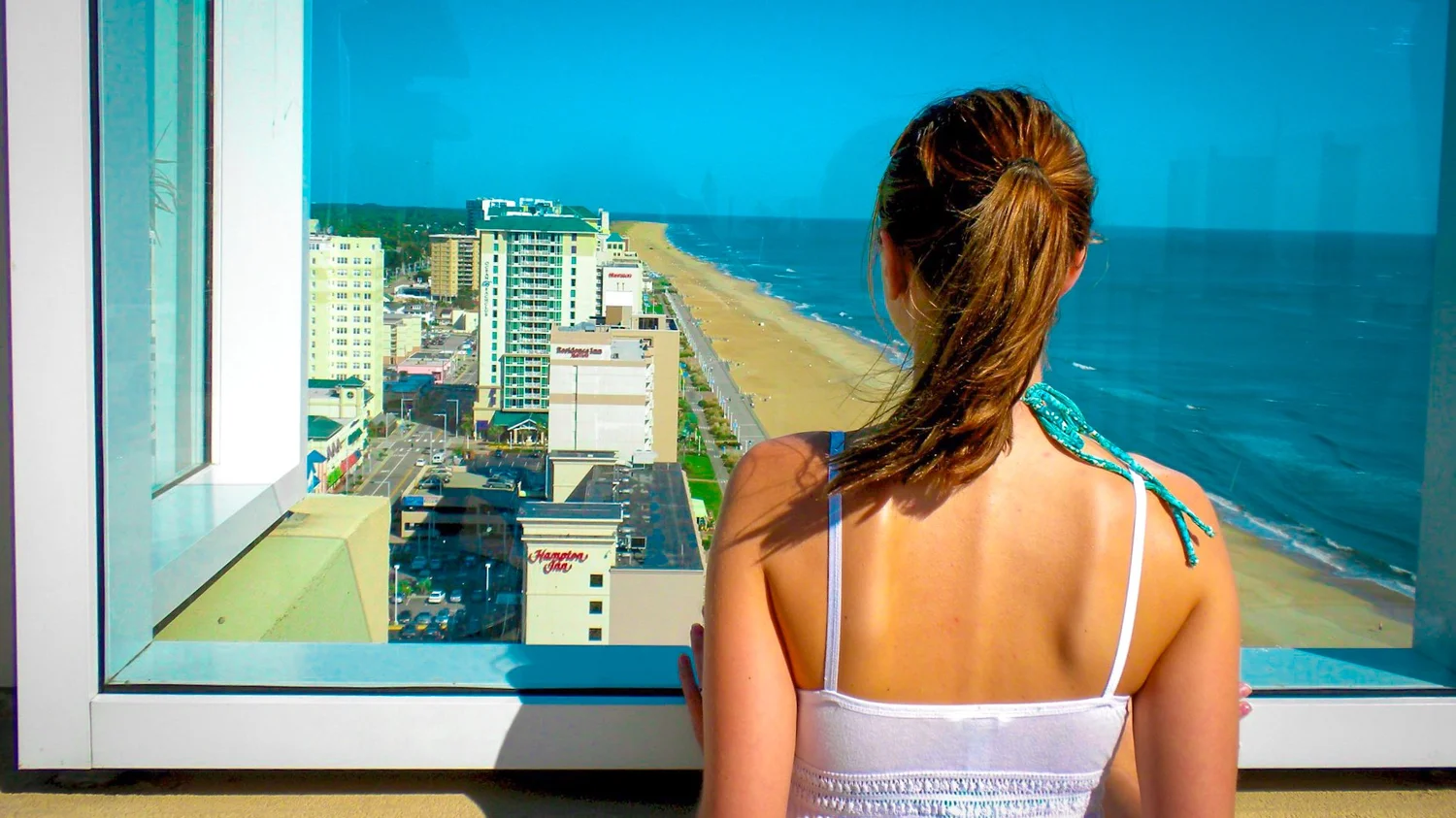 Woman seen from behind looking out a hotel window at a beach and ocean